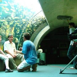Child and older male sit in front of aquarium fish tank in aquarium tunnel with director providing instructions in front of RED Epic Camera with Boom Mic Operator Standing besides camera tripod